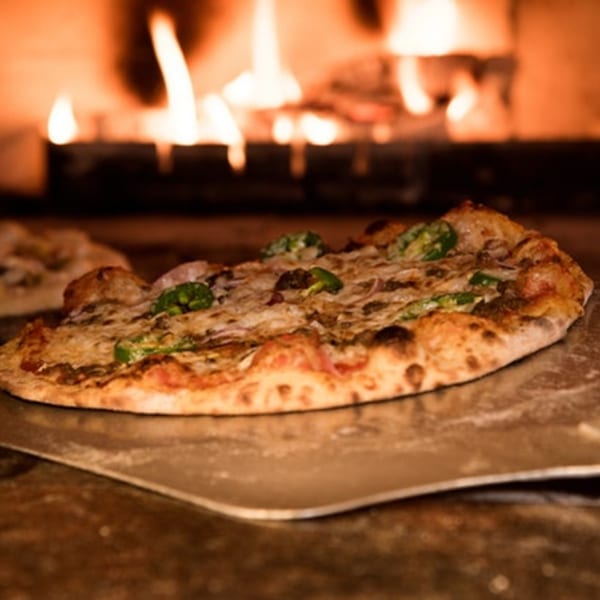 A freshly baked pizza with green peppers and cheese on a metal peel, in front of a wood-fired oven.