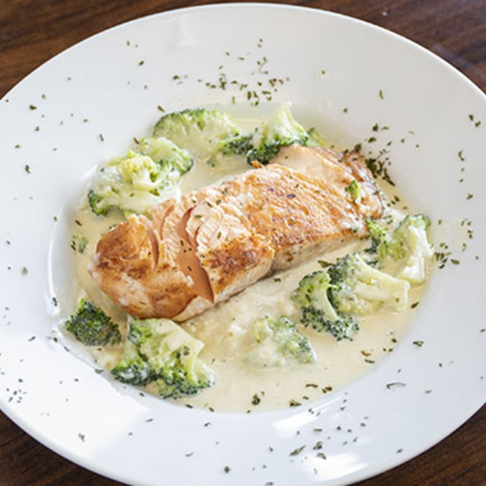 A salmon fillet with broccoli florets in a creamy white sauce, garnished with herbs on a white plate.