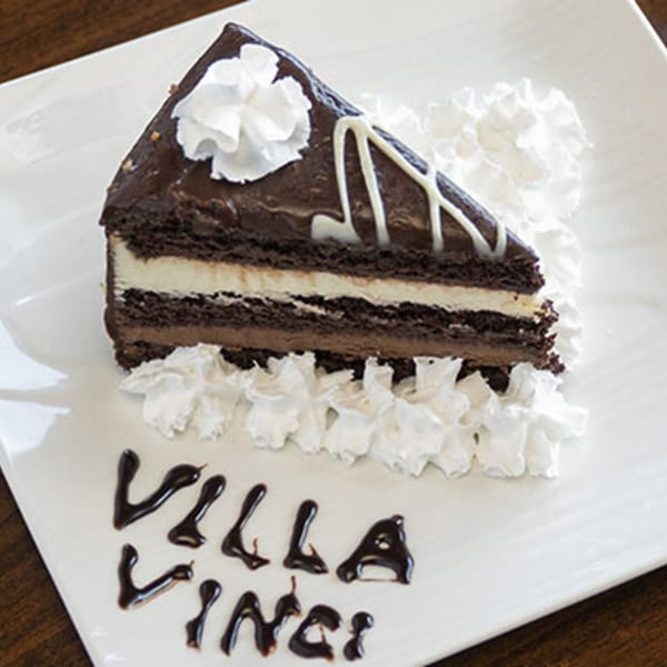 A slice of chocolate cake with white frosting layers, whipped cream, and "Villa Vinci" written in chocolate.